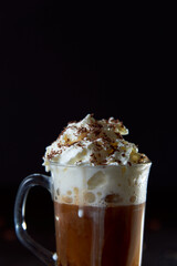 Close up of a glass cup of coffee with whipped cream and chocolate on it, chocolate chip cookies and roasted coffee beans on dark background. Concept of ready to eat food, tasty snack. Selective focus