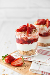 Homemade layered dessert with fresh strawberries, cream cheese or yogurt, granola and strawberry jam in glasses on white wood background. Healthy organic breakfast or snack concept. Selective focus.