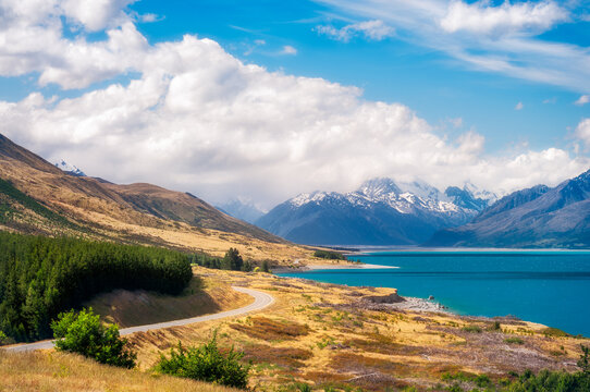 The spectacular winding road along the shore of Lake Pukaki, a glacial alpine lake in Mackenzie Basin in New Zealand's South Island, on a summer day with snow-capped mountain in fog in the distance.