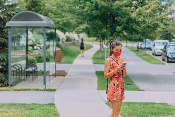 Mask wearing woman waiting for the bus holding mobile phone COVID-19 contact tracing app when using public transport. Obligatory face covering wear in many cities as corona virus prevention.