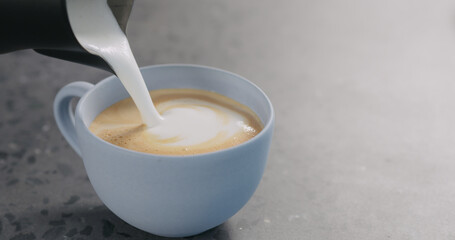 Pour steamed milk into espresso to make cappuccino in blue cup on concrete background