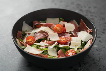 Salad with prosciutto, tomato, arugula leaves and parmesan cheese in black bowl on concrete surface