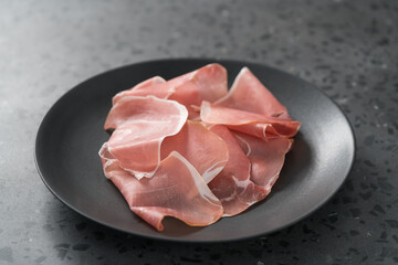 Thin italian prosciutto slices on black plate on conctere background