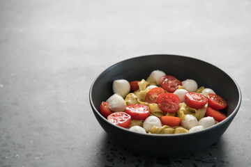 fettuccine pasta with pesto, mozzarella and cherry tomatoes in black bowl on concrete background with copy space