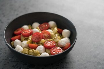 fettuccine pasta with pesto, mozzarella and cherry tomatoes in black bowl on concrete background with copy space
