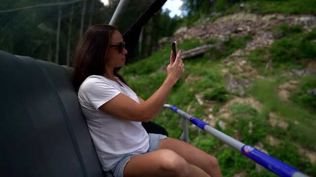 tourist woman is riding on cable railway and photographing by smartphone at summer day