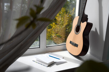 A yellow-brown acoustic guitar stands near the window. Nearby lies a notebook with a pencil