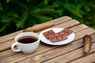 cup of coffee and chocolate bar on wooden table with spruce branches on background