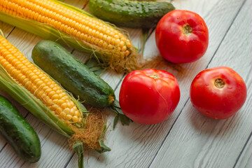 Ripe ears of corn, red tomatoes and fresh cucumbers lie on a light wooden table; vegetable bouquet, harvest