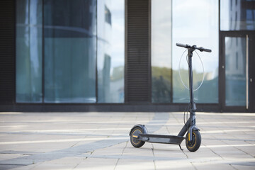 Background image of black electric scooter standing on tiled floor against glass building in urban...