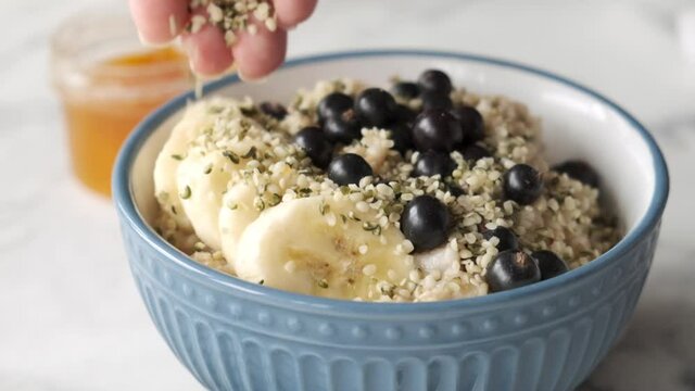 Slow motion adding hemp seeds into oatmeal porridge bowl with bananasand black currants. Healthy breakfast, vegan food concept