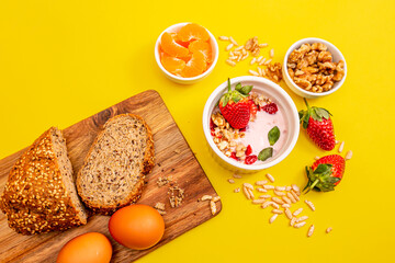 Healthy breakfast with granola, eggs, homemade bread,  and fruits on a wooden table, in white bowls, with a yellow background. 
