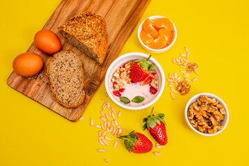 Healthy breakfast with granola, eggs, homemade bread,  and fruits on a wooden table, in white bowls, with a yellow background. 