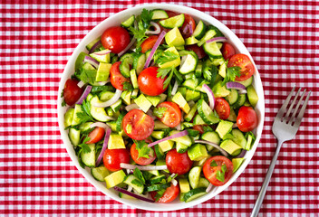 Salad of fresh ripe tomato, onion, avocado, cucumber and parsley and olive oil, salad in a white plate with a fork and towel on a red checkered tablecloth