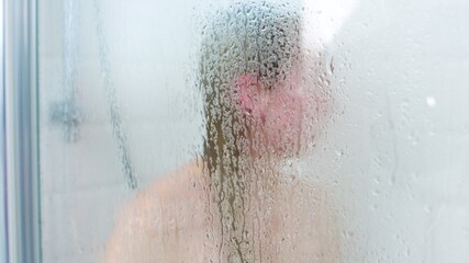 Unrecognizable woman standing in shower cabin behind steamy glass with waterdrop.
