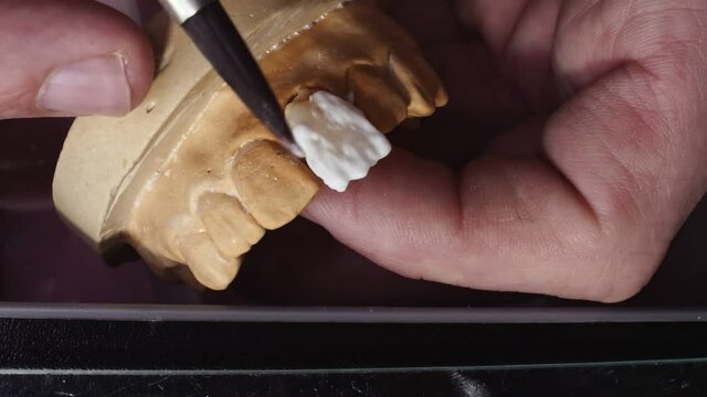 Close up view of dental technician draws white ceramics glaze on prosthesis of teeth in workplace in laboratory of implants
