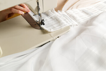 A woman works on a sewing machine. seamstress sews white curtains, close up view.
