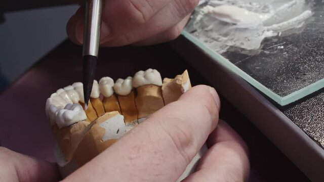 Close up view of dental technician draws white ceramics glaze on prosthesis of teeth in workplace in laboratory of implants