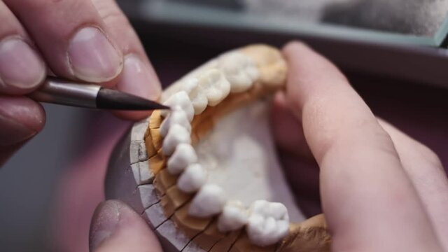 Close up view of dental technician draws white ceramics glaze on prosthesis of teeth in workplace in laboratory of implants