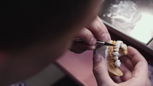 Close up view of dental technician draws white ceramics glaze on prosthesis of teeth in workplace in laboratory of implants