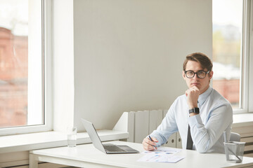 Minimal portrait of young businessman wearing glasses and looking at camera while sitting at desk in white office interior, copy space