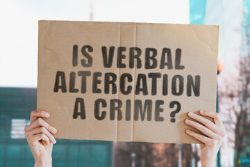 The question " Is verbal altercation a crime? " on a banner in men's hand with blurred background. Conflict. Street. Communication. Relationship. Confrontation