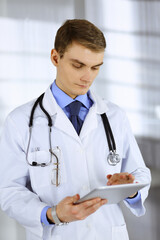 Young doctor man standing at his hospital office, with a stethoscope, using a computer tablet. Perfect medical service in clinic. Happy future in medicine and healthcare