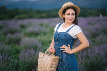 Portrait of attractive woman in lavender field in straw hat and denim sundress. Farm style in lavender with straw bag