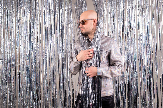 Strange caucasian man in a glittering white shirt, black leather pants, grey shoes and black sunglasses poses with shiny silver tinsel