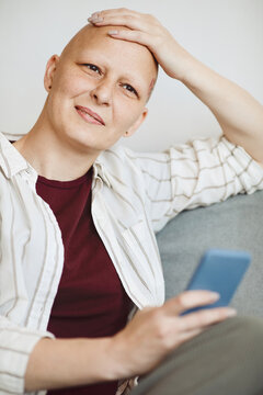 Vertical portrait of smiling bald woman rubbing head and looking away pensively while sitting in cozy armchair at home, alopecia and cancer awareness
