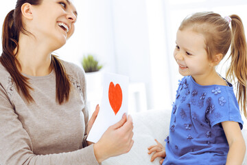 Mother's day concept. Child daughter congratulates mom and gives her postcard with red heart shape. Mum and girl happy smiling and hugging. Family fun and holiday
