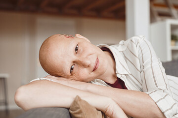 Minimal close up portrait of bald adult woman smiling at camera while lounging on couch in...