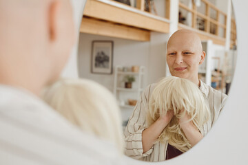 Reflection portrait of bald adult woman looking in mirror holding wig while standing in warm-toned...