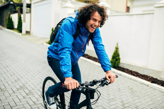 Image of handsome cyclist man cycling on his bike down the street next to the house. Caucasian male courier with curly hair delivers parcel cycling with a bicycle in the city in a rainy day