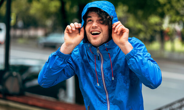 Image Of Man Smiling Broadly, Wearing Blue Raincoat During The Rain Outside. Handsome Happy Male In Blue Raincoat Enjoying The Rain In The City Street. The Guy Has Joyful Expression In Rainy Weather.