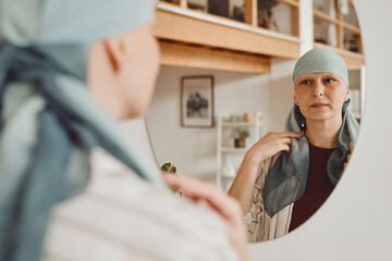 Warm-toned portrait of modern bald woman putting on head scarf while looking in mirror standing in...