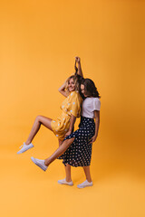 Enthusiastic female friends in trendy gumshoes funny dancing on yellow background. Wonderful...