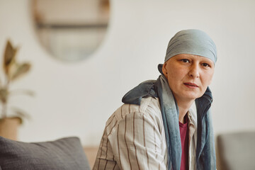 Warm-toned portrait of bald adult woman wearing headscarf looking at camera while posing in minimal...