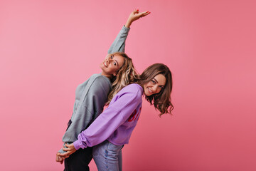 Blissful female friends in street clothes holding hands on pink background. Positive caucasian ladies dancing with pleasure.