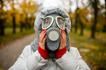 Picture of strange female in a warm white jacket with a gas mask on her head goes for a walk in the autumn park