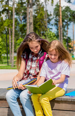 Two schoolgirls are sitting on a bench in the park, doing homework