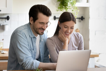 Fototapeta premium Happy young husband and wife sit relax in modern home kitchen look at laptop screen shopping online, smiling millennial Caucasian couple rest on weekend browse surf wireless internet on computer