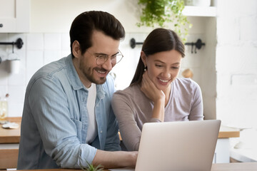 Happy young husband and wife sit relax in modern home kitchen look at laptop screen shopping online, smiling millennial Caucasian couple rest on weekend browse surf wireless internet on computer