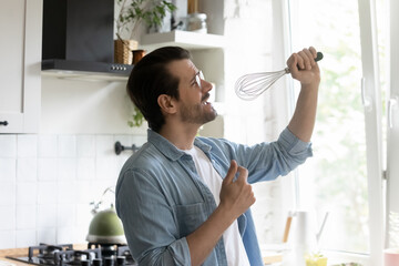 Overjoyed young Caucasian man enjoy lazy leisure weekend at home sing in kitchen appliance, happy millennial male have fun cooking at home, go crazy engaged in funny entertainment activity