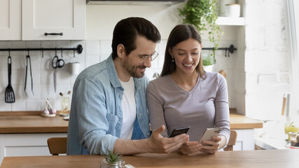 Smiling millennial couple sit at kitchen using modern smartphone gadgets together, happy young Caucasian man and woman spouses browsing surfing wireless internet on new cellphone devices