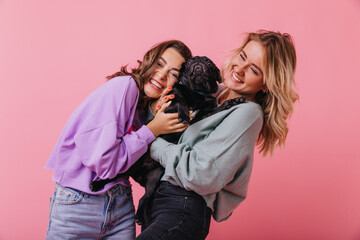 Pretty girls in casual outfit laughing while posing with cute black puppy. Indoor shot of best friends holding french bulldog.