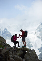 Travelling couple hiking in Swiss Alps, man helps woman to climb up, mount Ober Gabelhorn in clouds is on background, concept of adventure, tourism, alpinism in Alps