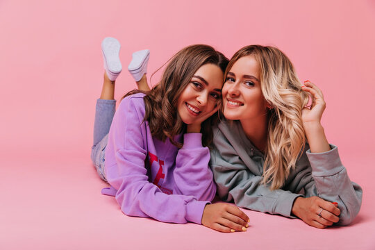 Curly blonde girl lying next to her best friend during photoshoot. Cute caucasian sisters posing on the floor and smiling.