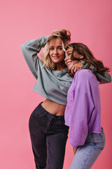 Sensual slim girl in black pants gently embracing her friend. Two attractive ladies in stylish casual clothes posing on pink background.