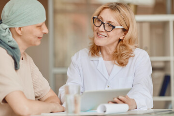 Warm-toned portrait of smiling female doctor talking to bald woman and showing data at digital tablet during consultation on alopecia and cancer recovery, copy space - obrazy, fototapety, plakaty